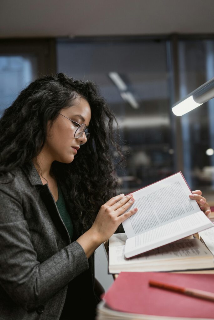 A woman with long curly hair and glasses reading a book at a desk under a lamp in a quiet study room.